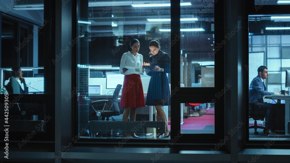 Poster shot from outside: two businesswomen working on a tablet computer in office. managers standing by th