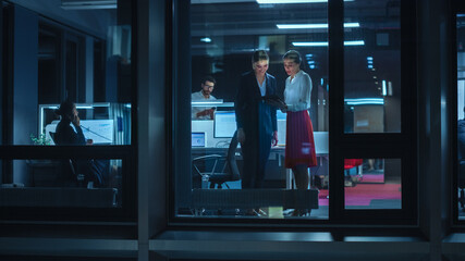 Fototapeta premium Shot from Outside: Two Businesswomen Working on a Tablet Computer in Office. Managers Standing by the Window. Employees Talk about Financial and Business Development. Colleagues Work Behind Them.