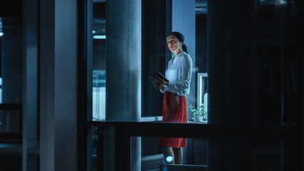 Fototapeta premium Shot from Outside: Businesswoman Working on a Tablet Computer in the Office. Manager Checking Emails while Standing by the Window. Employee Planning Financial Tasks and Business Development.