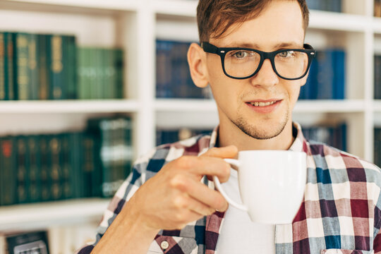 student in glasses and a plaid shirt drinks coffee and relaxes sitting at the table against the background of the wall with books