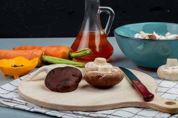 side view of fresh mushrooms with kitchen knife on a wooden cutting board and fresh carrots with a bottle of oil on light blue background