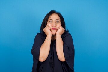 middle aged Arab woman standing against blue background with surprised expression keeps hands under chin keeps lips folded makes funny grimace