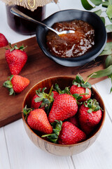side view of fresh ripe strawberries in a wooden bowl and jam in a saucer on wooden board