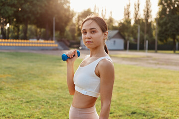 Young adult female with pleasant appearance wearing white sport top working out biceps and triceps, using blue dumbbell for training outdoor in stadium.