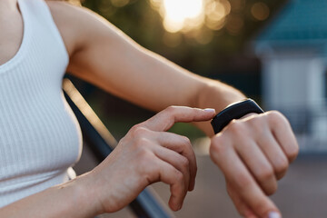 Faceless woman checking fitness and health tracking wearable device on her hand, unknown female in white top posing outdoor in stadium on sunset.