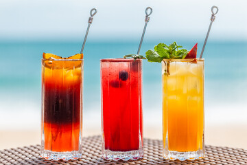 Close-up of Set of Three Refreshing Fruit Cocktails Standing on Table on Beach, Blurred Turquoise Sea on Background. Colorful Summer Ice Cold Drinks. Fresh Healthy Beverages with Berries and Orange