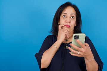 middle aged Arab woman standing against blue background thinks deeply about something, uses modern mobile phone, tries to made up good message, keeps index finger near lips.