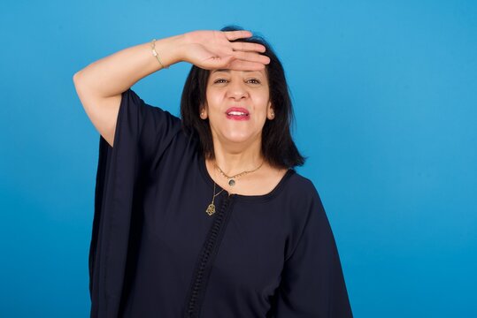 middle aged Arab woman standing against blue background wiping forehead with hand making phew gesture, expressing relief feels happy that he prevented huge disaster. It was close enough
