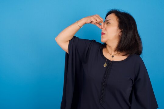 Displeased Middle Aged Arab Woman Standing Against Blue Background Plugs Nose As Smells Something Stink And Unpleasant, Feels Aversion, Hates Disgusting Scent.