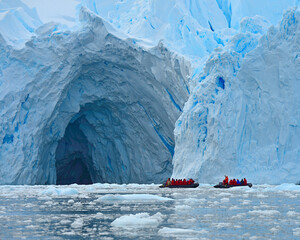 Two zodiac vessels explore the entrance of a large ice cave at the edge of an enormous glacier -...