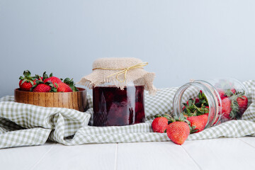side view of strawberry jam in a glass jar with fresh ripe strawberries on plaid fabric on white background