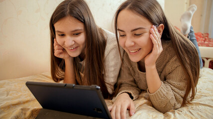 Portrait of two smiling teenage girls having video conference on tablet computer while lying on bed.