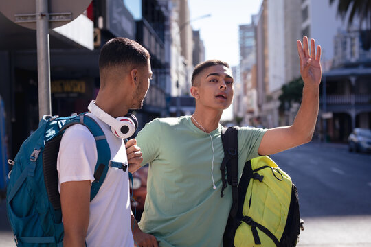 Two mixed race male friends with backpacks standing in sunny city street, one hailing taxi