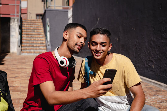 Two smiling mixed race male friends sitting in sunny city street taking selfie with smartphone