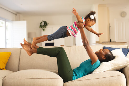 Happy african american mother lying on couch having fun balancing smiling daughter above her