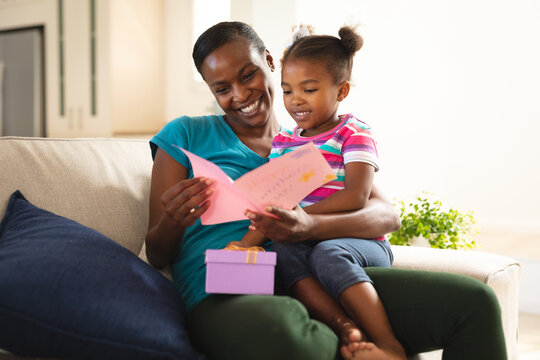 Happy african american mother and daughter sitting on couch with card and present from daughter