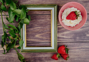 top view of an empty picture frame with fresh ripe strawberries cottage cheese in a bowl and green leaves on wooden background with copy space