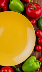 top view of an empty yellow plate and fresh vegetables colorful bell peppers tomatoes and cucumbers on wooden rustic background