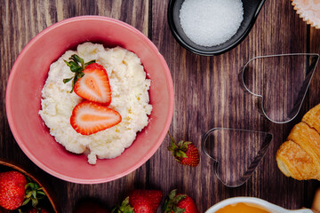 top view of cottage cheese in a pink bowl with fresh ripe strawberries and cookie cutters on rustic wooden background