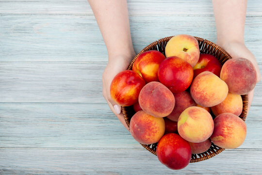 top view of female hands holding a wicker basket with fresh ripe nectarines and peaches at rustic wooden background with copy space