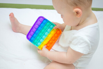 Little baby girl sitting at home on the bed, playing Pop-it new anti-stress toy, popular with children. Kid development idea.