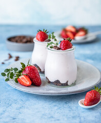 Two jars with yoghurt, chocolate granola and fresh strawberries on ceramic plate, blue concrete background.