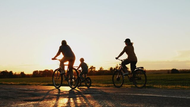 Mother with her teenage daughter and little son traveling on bicycles on a country road at sunset