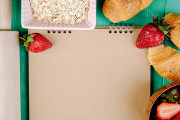 top view of fresh ripe strawberries on sketchbook and oatmeal flakes with croissants on green background