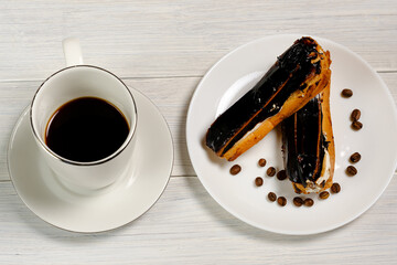 A white cup of coffee and two eclairs on a white plate with coffee beans