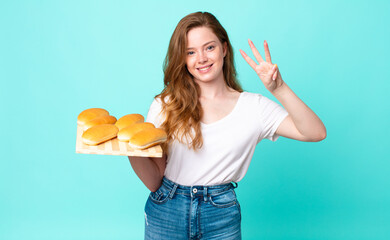 red head pretty woman smiling and looking friendly, showing number three and holding a bread buns...