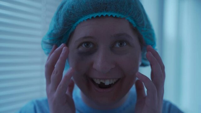 Mad woman with mental illness with dental injuries and facial batters in hospital gown looks somewhere and laughs in hospital. Personality disorder. Female patient having mental health problem.