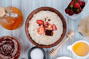 top view of oatmeal porridge in a wooden bowl with  fresh ripe strawberries and honey with jam on white wooden background