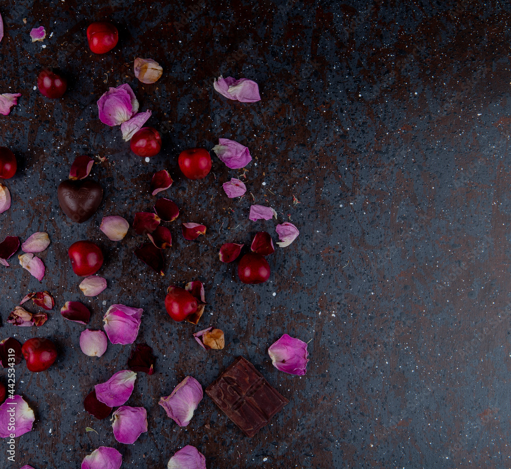 Poster top view of rose petals with heart shaped chocolate candies and red cherries on black background wit