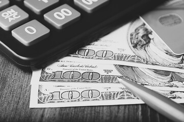 Dollars in one-hundred-dollar bills are spread out on the table next to a calculator, a credit card, and a pen. One-hundred-dollar bills. US dollars. Black and white style.