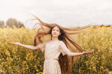 Young beautiful girl with long hair flying in the wind against the background of rapeseed field....