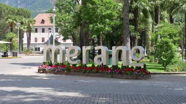 Merano, Italy, June 2021. In the public gardens in the center, the name of the city made of gray metal with brightly colored flowers at the base. In the background tourists.