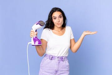 hispanic pretty woman feeling puzzled and confused and doubting and holding an iron steam
