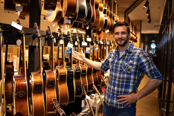Portrait of professional seller standing in music shop and showing collection of guitars for sale.