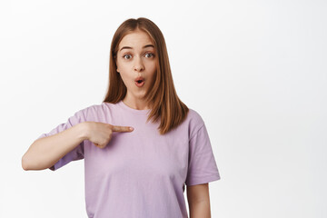 Fototapeta premium Portrait of young woman looks surprised, points at herself with amazed hopeful face, being chosen, winning smth, unexpected good news, standing against white background