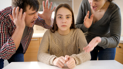 Father and mother shouting and screaming at teenage daughter sitting behind table on kitchen. Family violence, conflicts and relationship problems - obrazy, fototapety, plakaty