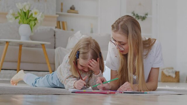 Caucasian mother babysitter wears glasses lies on floor of house with little daughter child preschool girl study together enjoy hobbies drawing picture draw with pencils artwork creative imagination