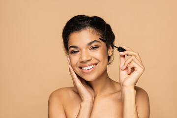 cheerful african american woman with bare shoulders applying mascara isolated on beige
