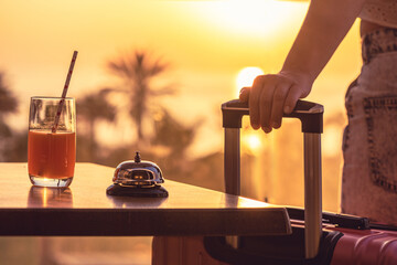 Woman with suitcase ringing hotel service bell with welcome drink and sea and palm tree view on sunset. Travel concept. 24-hour beach hotel front desk. Late check-out. - obrazy, fototapety, plakaty