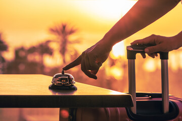 Woman with suitcase ringing hotel service bell with sea and palm tree view on sunset. Travel...