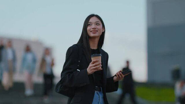 Happy brunette mixed-race young asian woman with coffee walking in the city, using smartphone and drinking hot takeaway coffee. Ethnicity. Urban lifestyle. - Powered by Adobe