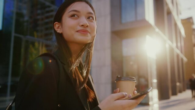 Inspired happy young asian mixed-race beautiful woman using smartphone, surfing internet on phone, enjoying morning sunshine outdoor. City. Modern people.