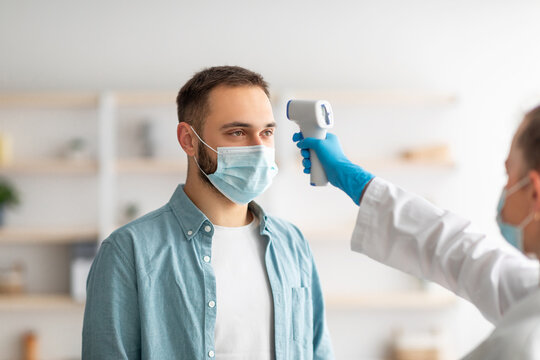 Doctor measuring young guy's temperature with electronic thermometer at home. Vaccination against covid concept
