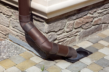 gutter made of brown pvc plastic drainage of rainwater from the roof of the house into the ground drainage system on the facade of the building lined with natural stone with a sidewalk made of tiles.