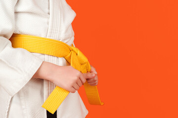 Kid in karate kimono holding his yellow belt, close up. Boy practicing karate on color background,...