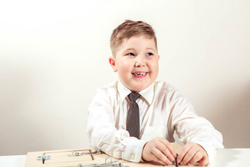 Naklejka premium little boy of 7-9 years old plays a board game, sits at a table in a light shirt against the background of a white wall in the room. Happy child studying at school